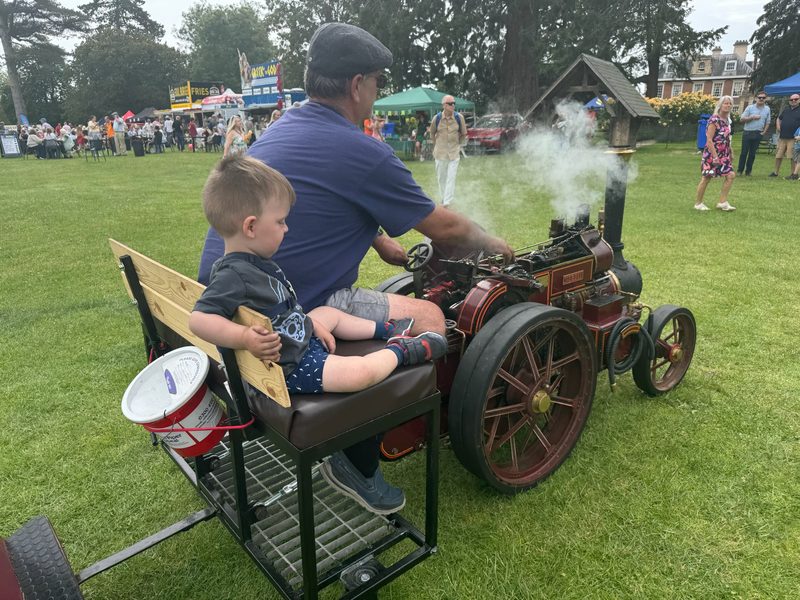 A young passenger rides Nellie's trailer at a busy summer fete, a charity collection bucket hanging from the seat rail