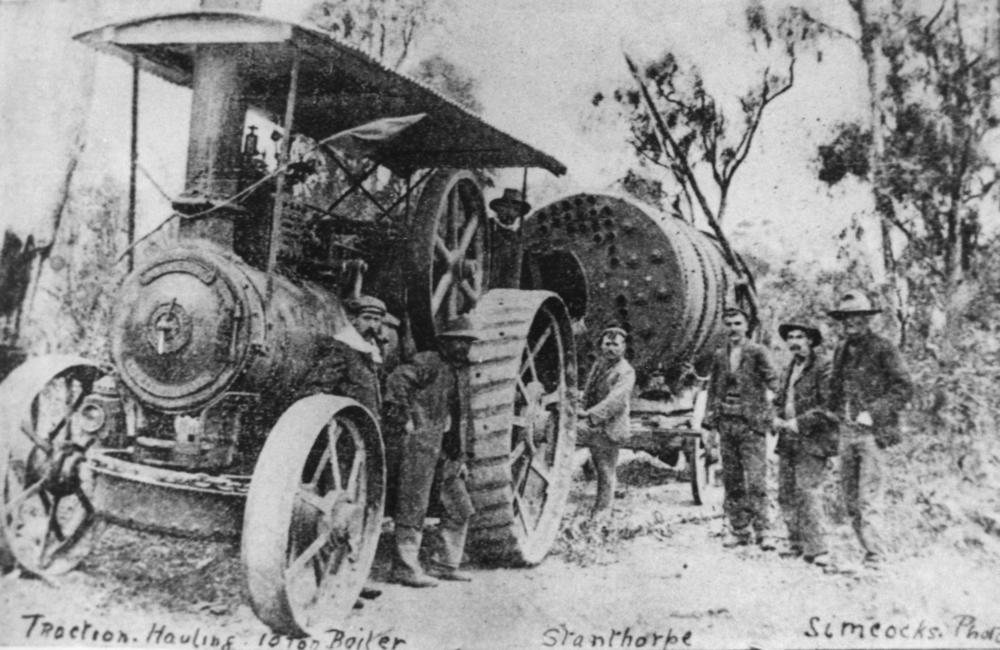 Black and white photograph from around 1910 of a traction engine hauling a ten-ton boiler along a dirt road, with workers standing alongside