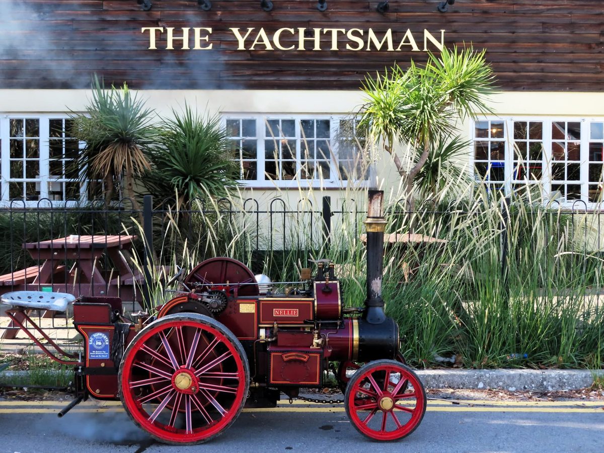 Side profile of Nellie in her Crimson Lake livery, parked outside The Yachtsman pub with steam drifting from her chimney